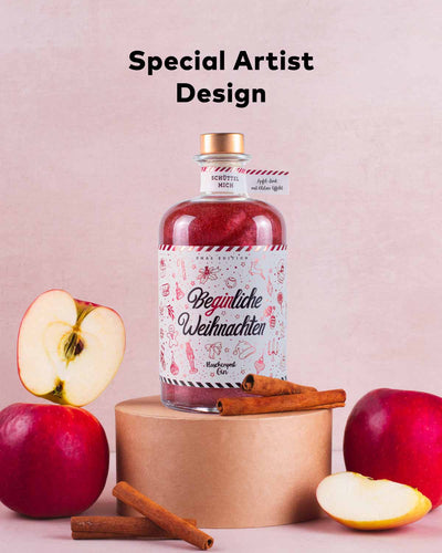 Die Beginliche Weihnachten Limited Edition steht auf einem Podest vor neutralem Hintergrund. Links und Rechts daneben sind Äpfel und Zimtstangen abgebildet. Es wird auf das Special Artist Design des Etiketts hingewiesen.